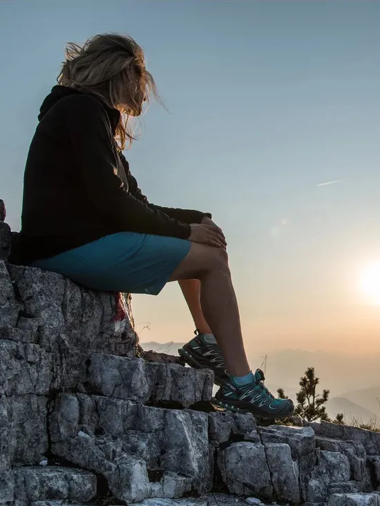Frau sitzt auf Felsen in den Bergen und blickt ins Tal bei Sonnenuntergang